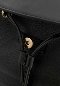 Gros plan sur un sac à main en cuir noir, montrant une surface texturée, des ferrures dorées, et une fermeture à bouton-pression détaillée avec un motif géométrique.