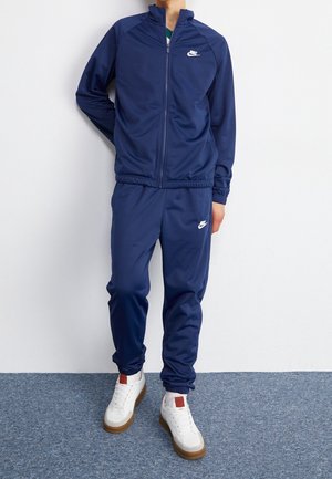 Personne portant un survêtement Nike bleu marine avec des logos blancs et des baskets blanches à semelles marron, debout devant un mur blanc et un tapis bleu.