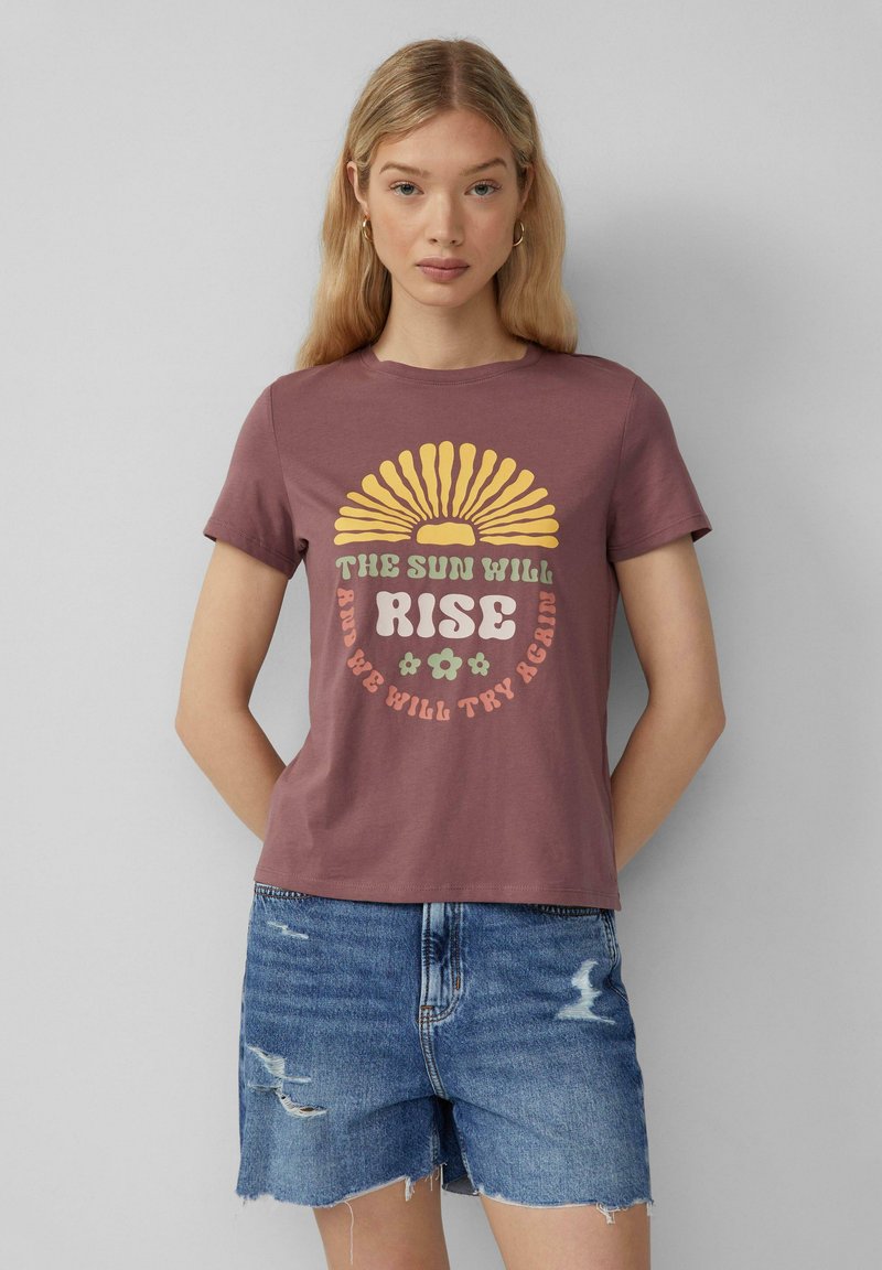 T-shirt en coton bordeaux avec un motif de soleil jaune et du texte en vert et rose. Assorti à un short en denim déchiré.