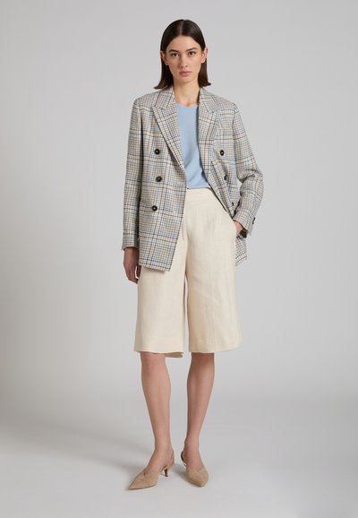 Vrouw staand, gekleed in een geruit dubbelbreasted blazer, lichtblauw topje, crèmekleurige knielange shorts en beige hakken, tegen een effen grijze achtergrond.
