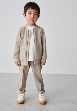 Beige Reißverschlussjacke mit langen Ärmeln, passende Hose und ein weißes T-Shirt darunter. Weiße Sneakers mit Klettverschlüssen und hellbraunen Sohlen.