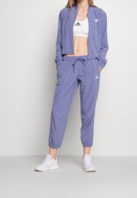 Ljuslila träningsset bestående av en cropped jacka och matchande joggers, tillverkat av mjukt material med elastiska muddar och logotyp på bröstet.