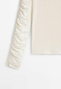 Langarmshirt in Creme mit geraffter Textur entlang der Ärmel, aus weichem Stoff gefertigt. Klassischer Schnitt mit einem schlichten Halsausschnitt.