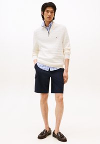 Cremfarbener Reißverschluss-Pullover mit gerippter Textur, kombiniert mit einem blau gestreiften Hemd mit Kragen und navyblauen Cargo-Shorts. Braune Slipper runden den Look ab.