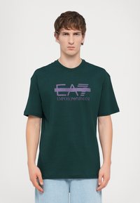 Zaļš kokvilnas T-krekls ar purpura krāsā drukātu logotipu "EA7 Emporio Armani" uz krūtīm. Īsas piedurknes un apaļais kakls.