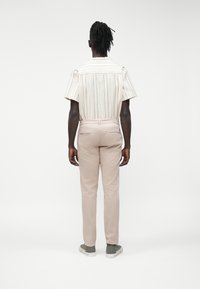 Mężczyzna z dredami stojący tyłem, ubrany w białą koszulkę z krótkim rękawem w pionowe paski, beżowe spodnie i szare trampki.