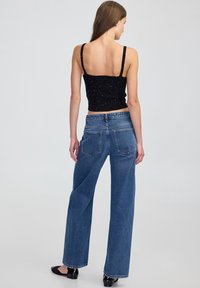 Žena oblečená v černém bezrukávovém topu a širokých modrých džínách, stojící bosky na světlém šedém pozadí, viděná zezadu.