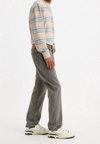 Homme portant un pull rayé beige et bleu avec un jean gris et des baskets blanches, debout devant un fond blanc uni.