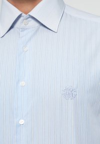 Jasnoniebieska koszula w paski na guziki z ostro zakończonym kołnierzykiem, z subtelnym haftowanym logo na piersi, gładka faktura materiału.