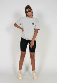 Light grey t-shirt with black logo on the chest, paired with black bike shorts and beige sneakers. Model wearing black sunglasses.