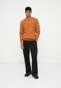 Πορτοκαλί πλεκτό πουλόβερ με υφές, ριμπ μανσέτες και στρίφωμα, συνδυασμένο με μαύρα παντελόνια σε ευρεία γραμμή και μαύρα παπούτσια χωρίς κορδόνια.