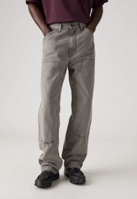 Pantalones de algodón grises con un corte relajado, que cuentan con detalles de costura visibles, bolsillos laterales y un cierre de botón, combinados con zapatillas deportivas negras.
