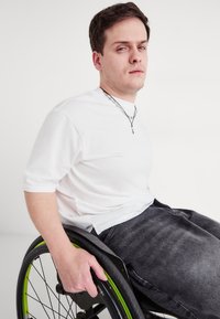 Biała koszulka krótki rękaw z okrągłym dekoltem, w zestawieniu z szarymi dżinsami. Osoba siedzi na czarnym wózku inwalidzkim z zielonymi akcentami.