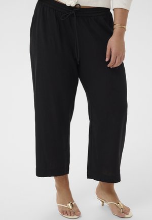 Kobieta ubrana w czarne, luźne spodnie o krótkiej długości z elastycznym pasem i sznurkiem, zestawione z beżowymi sandałami na niskim obcasie i złotymi bransoletkami.