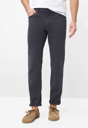 Mand iført mørkegrå bukser med opsmøg, beige snørede fritidssko og en hvid synlig skjorteærmekant, står med den ene hånd i lommen.