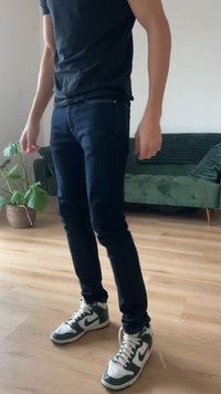 Ciemnoniebieskie jeansy o dopasowanym kroju, posiadające pięć kieszeni i subtelne przeszycia. Noszone z zielono-białymi wysokimi tenisówkami, stojąc na drewnianej podłodze.