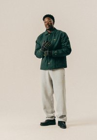 Ανδρας με σκούρο πράσινο σακάκι, γκρι φόρμες, μαύρες μπότες και καρό γάντια, στέκεται μπροστά σε απλή μπεζ βαφή.