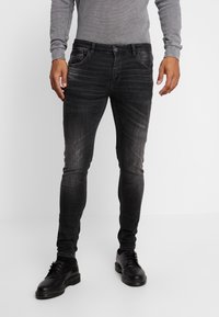 Jeans slim fit de color negro, hechos de denim, con un diseño desgastado, cinco bolsillos y cierre de botón, combinados con zapatos de cuero negros.