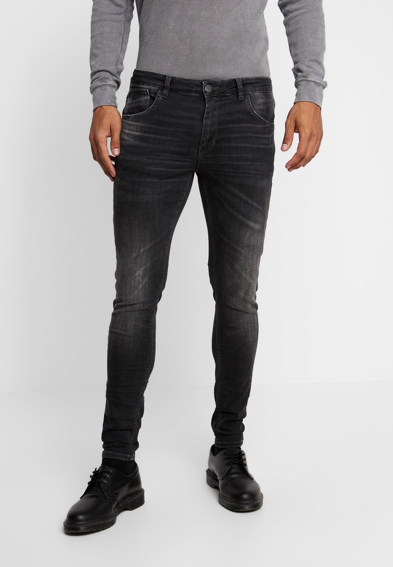 Jeans slim fit de color negro, hechos de denim, con un diseño desgastado, cinco bolsillos y cierre de botón, combinados con zapatos de cuero negros.