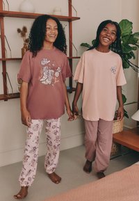 Dwie dziewczyny trzymają się za ręce, obie w kolorowych, za dużych t-shirtach. Jedna ma fioletowy t-shirt z grafiką kowbojskiego buta, druga jasnoróżowy. Spodnie mają wzory — jedne kwiatowe na jasnoróżowym tle, drugie z bordowymi paskami.