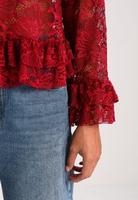 Rød blonde top med blomster mønster, flæseærmer og struktureret finish, parret med denim jeans. Nærbillede af ærmedetalje og hånd.