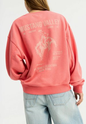 Osoba nosząca wyblakłą koralową bluzę z "Mustang Valley" i grafiką zawodnika rodeo, w połączeniu z jasnoniebieskimi dżinsami.