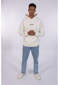 Crème hoodie met de tekst "crystal" in het zwart, voorzak, gecombineerd met lichtblauwe jeans en witte sneakers. Eenvoudig en modern ontwerp.