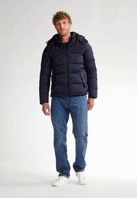 Marinblå vadderad jacka med dragkedja framtill, flera fickor och avtagbar huva, kombinerad med blå jeans och vita sneakers.