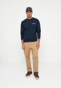 Moški stoji obrnjeni naprej, oblečen v temno modro majico s kapuco, svetle bež hlače, temno rjave teniske in temen klobuk pred enobarvno belo ozadjem.