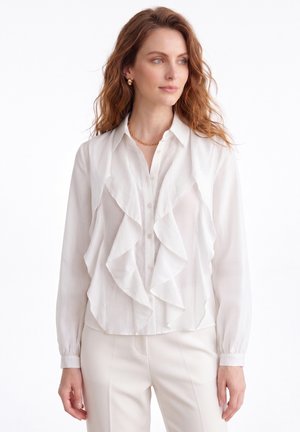 Femme aux cheveux châtain clair portant un chemisier blanc à volants, un pantalon blanc, un collier chaîne doré et des boucles d'oreilles en anneau doré, regardant sur le côté.