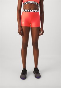 Røde treningeshortser med en svart "Under Armour"-livlinning, laget av et glatt, elastisk materiale. Bærer mørke, teksturerte sneakers.