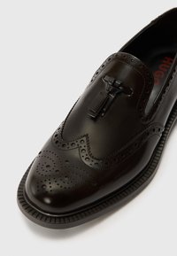 Black leather shoe with perforated brogue detailing, sleek shape, and a flat strap accent near the top. Textured sole with grooves.