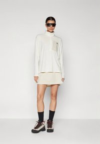 Fehér, hosszú ujjú, cipzáras galléros ing, mellkasi zsebbel, bézs szoknyával párosítva. A modell fekete napszemüveget és túracipőt visel.