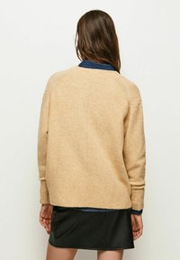 Pull en tricot beige avec ourlet et poignets côtelés, présentant un col rond et une coupe décontractée, porté par-dessus une chemise à col bleu foncé.