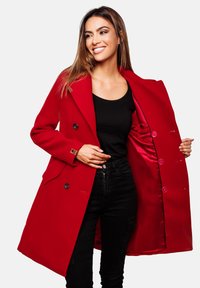 Roter Wollmantel mit doppelt geknöpften Knöpfen, breiten Revers und seitlichen Taschen. Mit einer Satin-Innenausstattung. Offen über einem schwarzen Oberteil getragen.