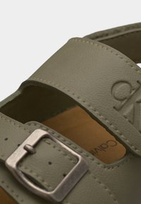 Zbliżenie na oliwkowozielony pasek skórzany sandała z metalową klamrą i tłoczonym logo Calvin Klein.