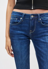 Niebieskie dżinsy z denimu o wąskim kroju, z pięcioma kieszeniami i żółtym przeszyciem. Tkanina ma gładką fakturę z lekkim przetarciem.