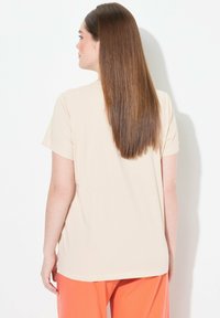 Camiseta de manga corta beige con corte recto. Tejido suave, escote redondo y sutiles aberturas laterales, mostrada desde la parte de atrás sobre un fondo blanco.
