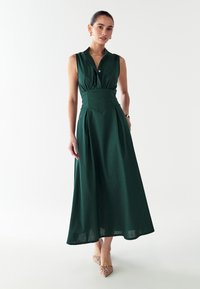 Grünes, ärmelloses Maxikleid mit einem knopfbaren Kragen, tailliertem Schnitt und plissiertem Rock. Getragen mit beigen spitzen Schuhen.