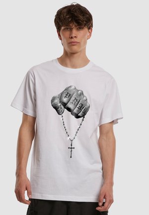 Junger Mann trägt ein weißes T-Shirt mit einer schwarz-weißen Grafik einer tätowierten Faust, die einen Rosenkranz mit Kreuz hält.