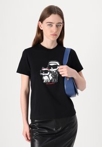 Svart bomullströja med grafiskt tryck av en katt och en figur, korta ärmar, med rund halsringning. Modellen håller en blå handväska.