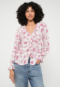 Blusa floreale con scollo arricciato, motivo a rose rosa su sfondo bianco, maniche lunghe con polsini abbottonati e orlo leggermente curvo.