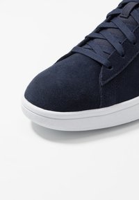 Zapatilla deportiva de ante azul marino con punta redonda, suela plana de goma blanca y cordones a juego. Presenta parte superior texturizada y detalles de costuras mínimas.