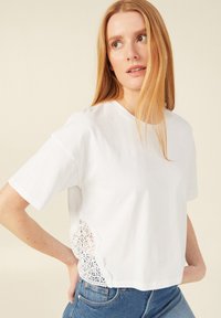 Witte cropped t-shirt van zacht katoen, met korte mouwen en een delicate kanten accent aan de linkerkant, gecombineerd met een blauwe jeans.