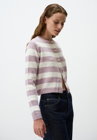 Cardigan all'uncinetto con strisce bianche e lilla, dotato di sei bottoni rotondi dorati e polsini a coste, abbinato a jeans blu scuro.