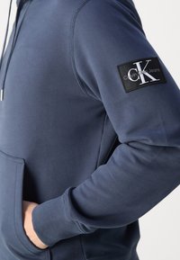 Felpa blu scuro con tasca frontale, cappuccio regolabile con coulisse e un'etichetta nera con "CK" sulla manica sinistra. Tessuto dal texture morbida.