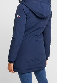 Navy blue padded parka with a hood, featuring ribbed cuffs and a logo patch on the sleeve. Smooth fabric with a slightly glossy finish.