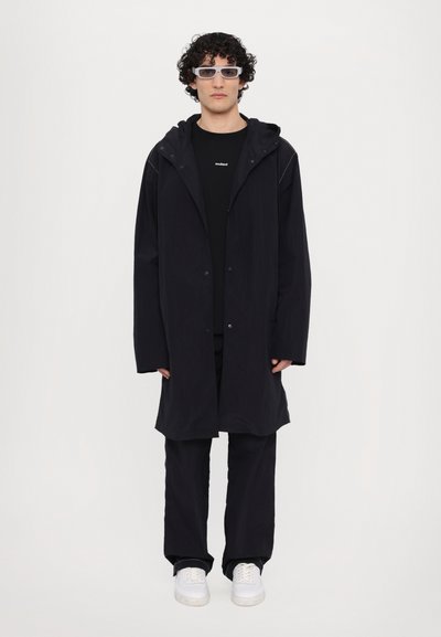 Zwarte hoodie met drukknoopsluitingen, gecombineerd met een zwart shirt en zwarte broek. Witte sneakers maken de look compleet. Model heeft krullend haar en draagt een bril.