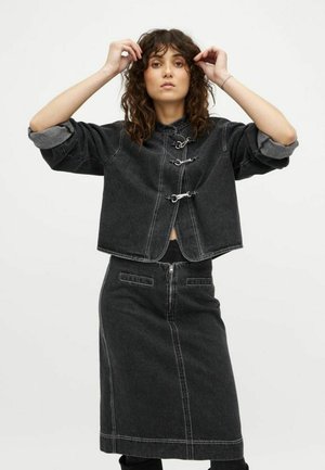 Woman with curly hair wearing a black denim jacket with metal clasps and matching high-waisted skirt, adjusting hair against a plain background.