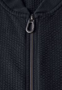 Pull noir avec une surface texturée, doté d'une fermeture éclair métallique et d'un col montant. La fermeture éclair est bordée d'un tissu contrastant.